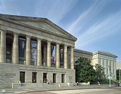 Large building with columns and landscaping. Blue sky in the background. Street in the foreground.