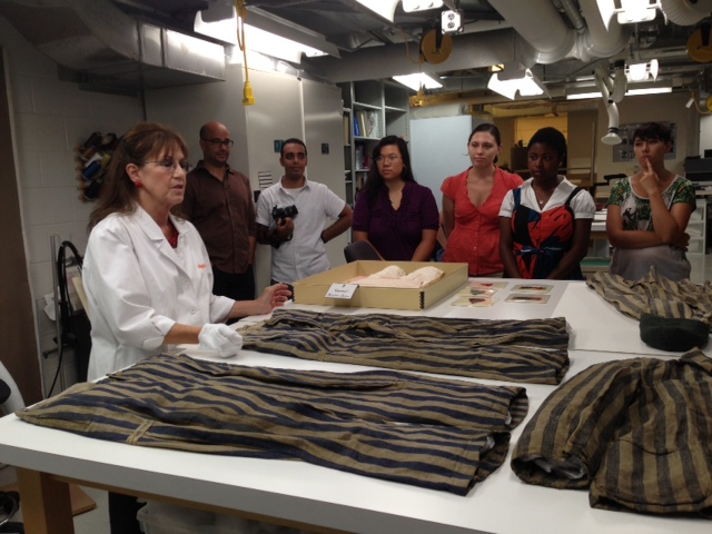 A group stands in front of a large table filled with historic artifacts. Conservator with gloved hands presents.