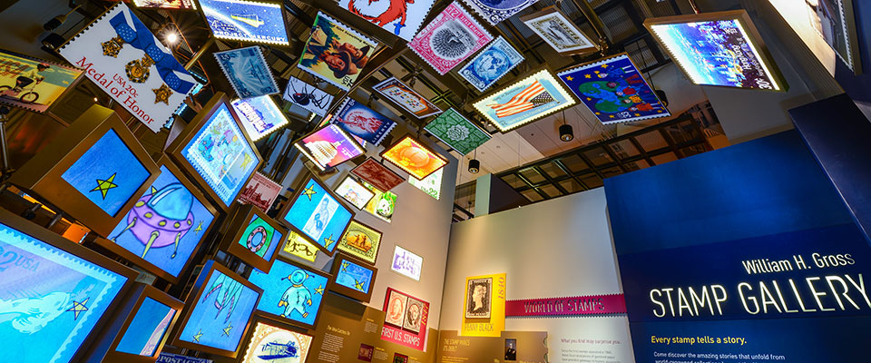 Colorful exhibit space featuring dozens of postal stamp designs filling the wall and covering the ceiling. Exhibit text reads the title of the Gallery