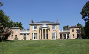 A reddish-tan stone, federalist façade with surrounding grounds on a sunny, cloudless day.