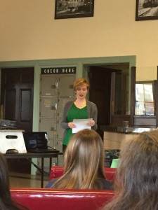 Speaker stands at the front of a crowded room, reading from a paper