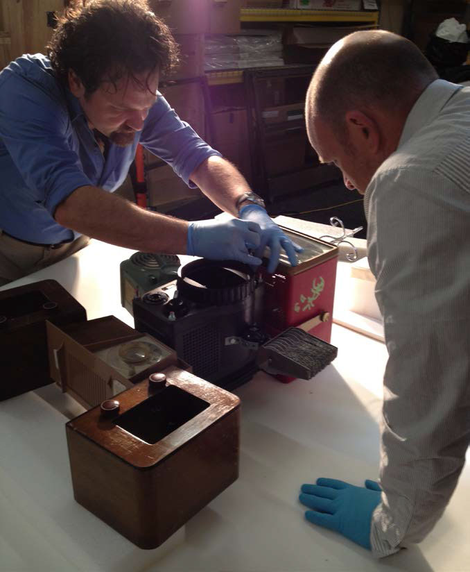 Two conservators wearing gloves work with an antique projector or radio