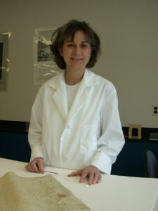 Woman in a lab coat and holding a metal spatula near an old document on a bench