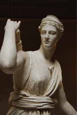 White stone-like sculpture of a greco-roman woman wearing tunics reaching for something from a basket on her right shoulder