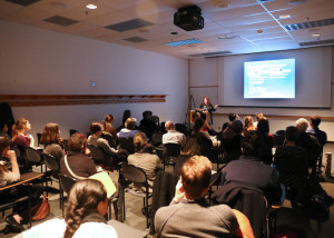 Speaker presents at podium in a darkened lecture room with presentation projected on screen behind.