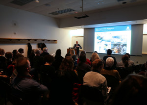 Speaker stands at podium in a darkened lecture room with presentation projected behind on wall.