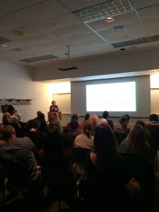 Speaker presents in a darkened lecture room with presentation projected on screen.