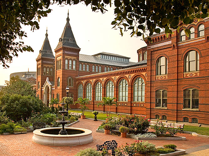Formal brick laid garden with flowering bushes, potted plants, and circular fountain. Garden sits in front of an ornate large brick building with two towers and large windows.