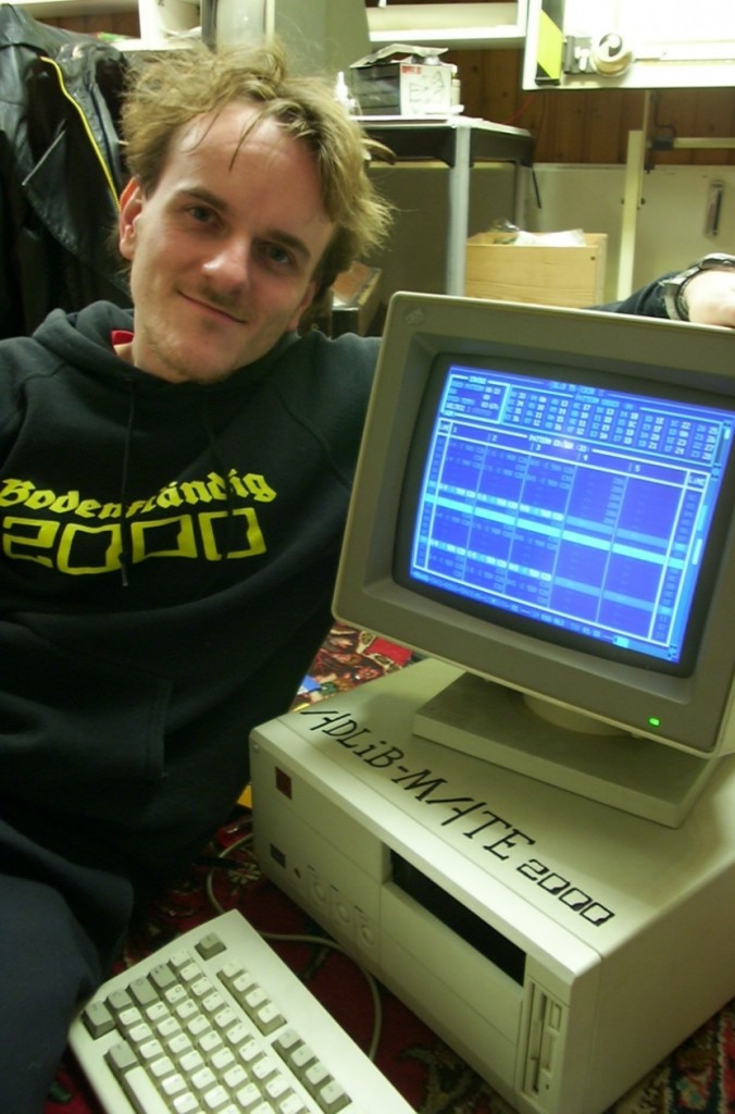 The speaker poses for a portrait half-hugging a vintage beige computer labeled as "ADLiB-MATE 2000". Illegible blue screen on the monitor.