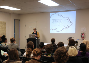 Speaker presents at the front of a lecture room with presentation projected on the wall behind.