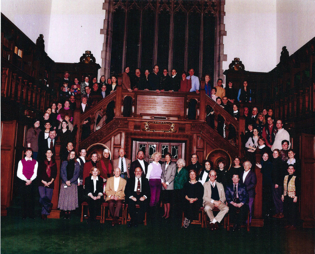 A large group of people stand in an ornate wood-paneled room up stairs and across a raised platfrom.