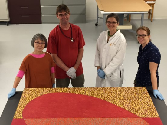 Four people stand in front of a large painting laid horizontally on a table in front of them