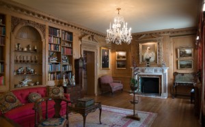 Ornate Victorian-era wood paneled room with portrait over the fireplace and bookshelves with books and small sculptures on display