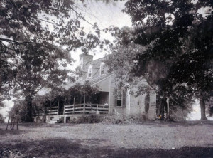 Black and white photo of a historic farm house with large trees in the yard. People sit on the porch posing for the camera.