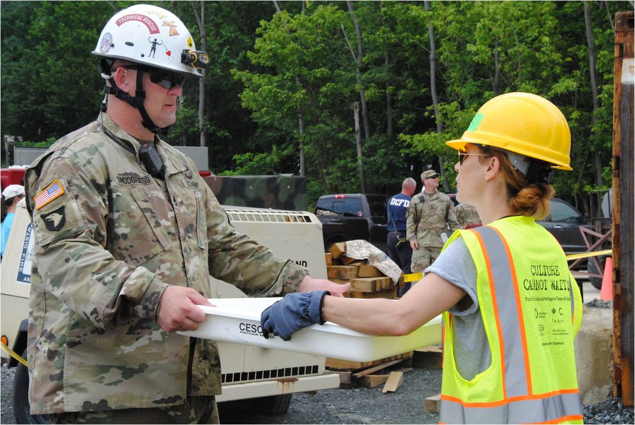 A workshop member in a hard hat and safety vest hands a shallow plastic tray to a member of the military in a hard hat