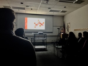 Speaker at podium presents in a dark room with presentation projected behind. An overexposed image of two red shoes with a small heel with round microscope images of condition details floating around the shoes.