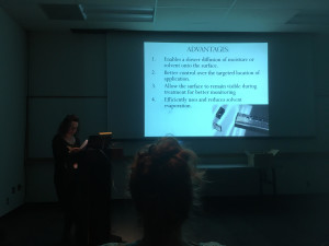 Speaker at podium presenting in a dark room. Presentation projected behind. Slide reads "Advantages: 1. Enables a slower diffusion of moisture or solvent onto the surface. 2. Better control over the targeted location of the application. 3. Allow the surface to remain visible during treatment for better monitoring. 4. Efficiently uses and reduces solvent evaporation.