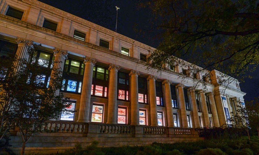 Large Federal building with columns at night. Lit windows feature stamps from all eras.