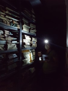 A worker wearing a dust mask and flashlight headlamp stands next to a full archives storage shelf in dark room