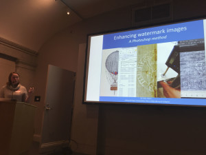 Speaker at podium in a dark room, presentation projected on screen behind. Presentation shows titled slide with photo collage of watermarks and spreadsheets