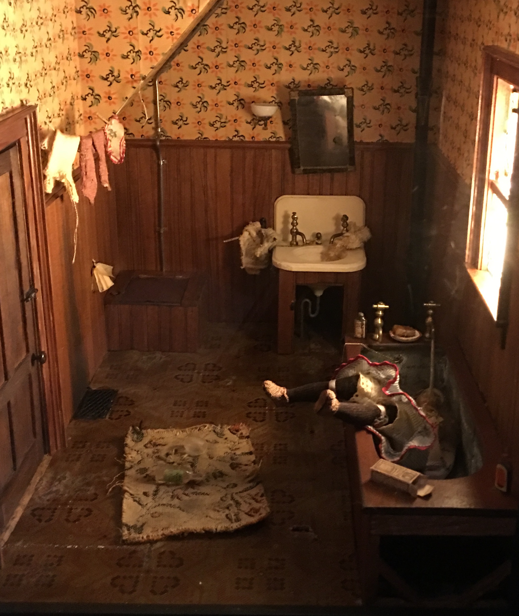 Closeup photo of a nutshell model as if the photographer is in the room. A wallpapered and wood-paneled bathroom with glass and bottle on floor. Doll in bathtub with legs projecting into air.