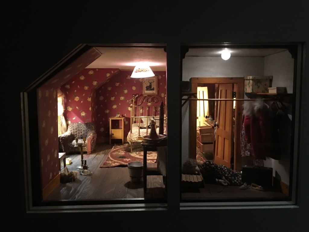 Closeup photo of a nutshell model as if the photographer is peering through a window or whole in the wall. A wallpapered bedroom with walk-in closet visible. Rooms filled with to-scale furniture and accessories. Doll face down in closet in foreground.