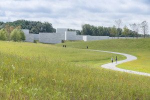 Grassy meadows surround a winding path to a blocky white building in the distance in front of a line of trees. People walk on the path toward the building.