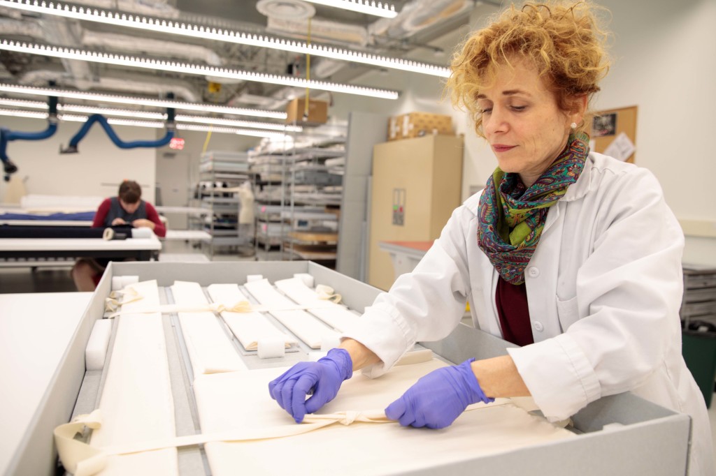 A conservator in a lab coat and gloves ties twill tape ties over artifacts in a blue board box.