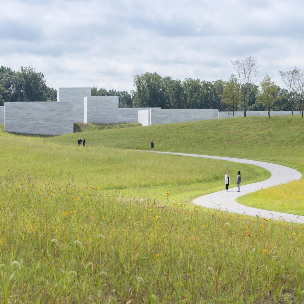 Grassy meadows surround a winding path to a blocky white building in the distance in front of a line of trees. People walk on the path toward the building.