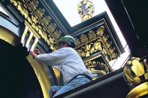 A worker in a hardhat working at the top of a ornately decorated and gilt archway and columns