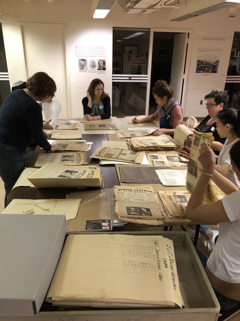 A group of seven people file through boxes of delicate yellowed newsprint, adding them to new acid-free folders. New metal-edged archival boxes sit in the foreground