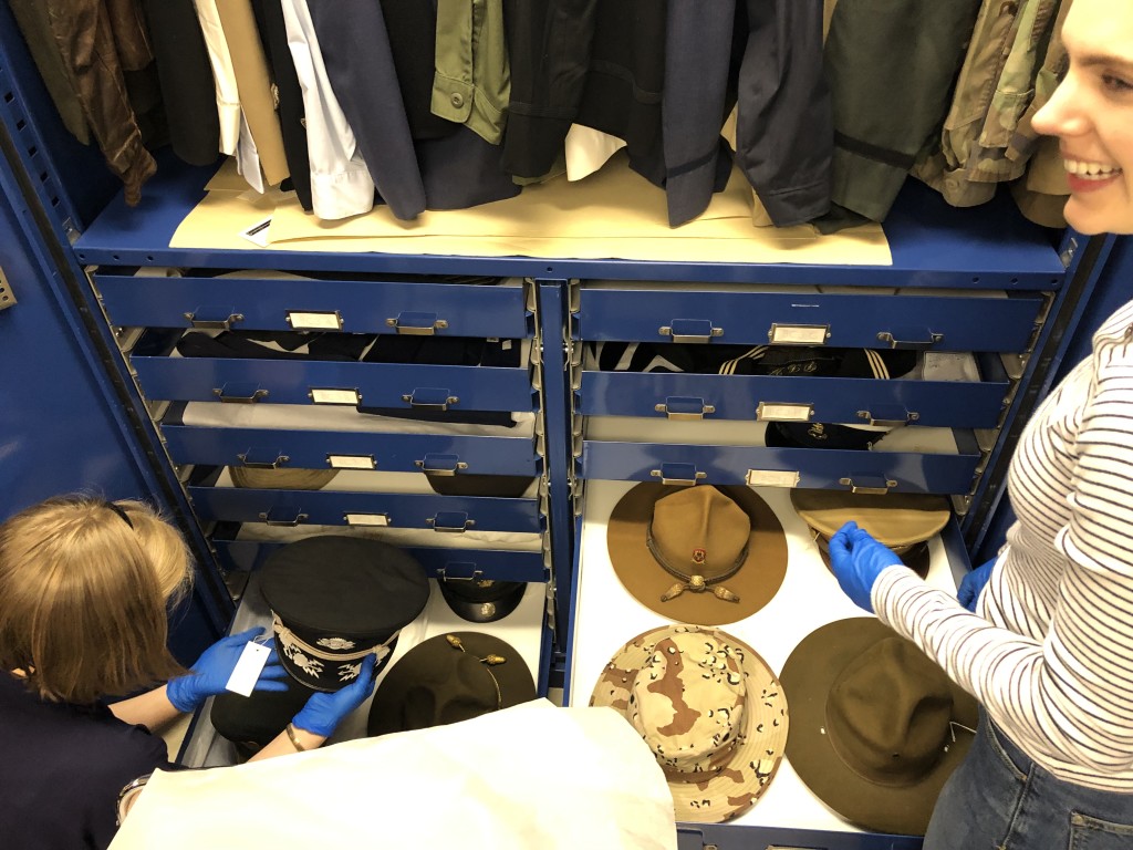 Two volunteers open blue metal drawers filled with a variety of hats