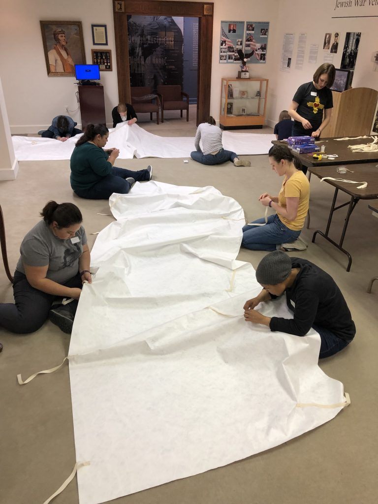 Nine volunteers sit on the floor as they sew straps onto large rolls of white Tyvek
