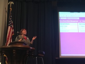 Speaker animatedly presents at ornate wood lectern with presentation displayed behind