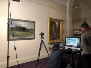 Two people look at a screen attached to a camera. Camera is on a tripod in front of a landscape painting in a gallery.