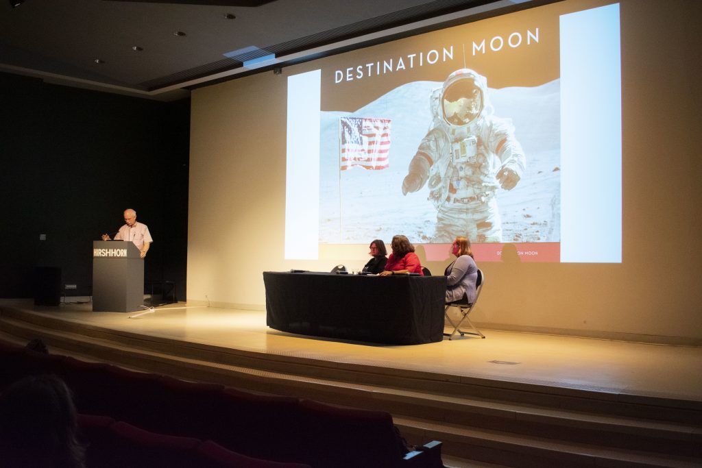 Panel of speakers on stage with presentation projected behind and above them