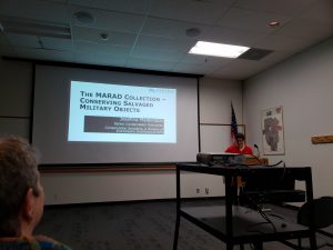 Speaker in a darkened lecture room with presentation projected behind them. Title slide showing