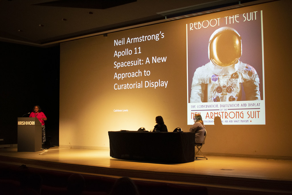 Panel of speakers on stage with presentation projected behind and above them