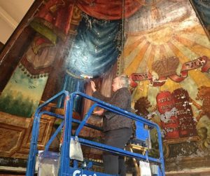 Conservator on a hydraulic lift holding a color card against a damaged mural in a curved, high space.