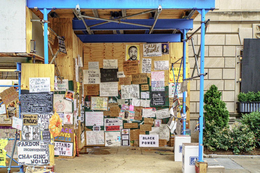 Scaffolding with handwritten signs, art, and pictures. Examples read Black Lives Matter; Pardon the inconvenience, changing the world; Know justice, know peace; #SayHerName; we will not rest until there is an end to racist policing; among other handwritten posters.