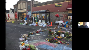 Screenshot of a virtual presentation. Speaker Jeanelle Austin presents an image of the George Floyd memorial. Floral bouquets, handwritten and printed signs, and other material surround colorful sidewalk chalk designs drawn on the street in front of a convenience store and sheltered bus stop.