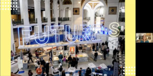 Screenshot of a virtual presentation. Oversized neon sign reading "FUTURES" with crowd of people wandering exhibit space below