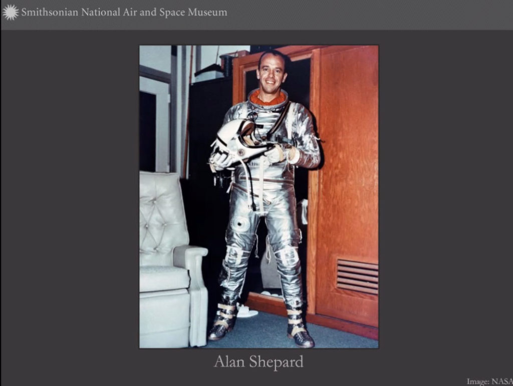 Screenshot of a virtual presentation. Photo of Alan Shepard wearing shiny silver-colored spacesuit holding helmet
