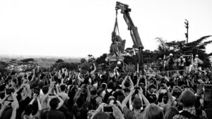 A crane crew lifts a statue among a crowd documenting with their phone cameras.