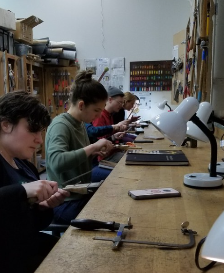 People seated at a workbench working with various tools to make silver rings.
