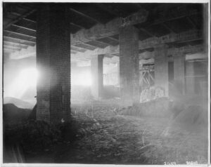 Black and white photo of empty, dusty room under construction. Bare brick columns and cement ceiling with piles of dirt and wheeled cart tracks in the dirt floor.