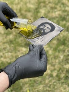 Person wearing black nitrile gloves pours a yellow liquid over a black and white photograph