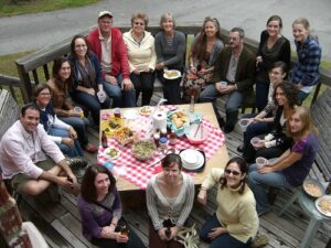 A group of 17 people circle around a table full of snacks on a wood deck. They smile at the camera.