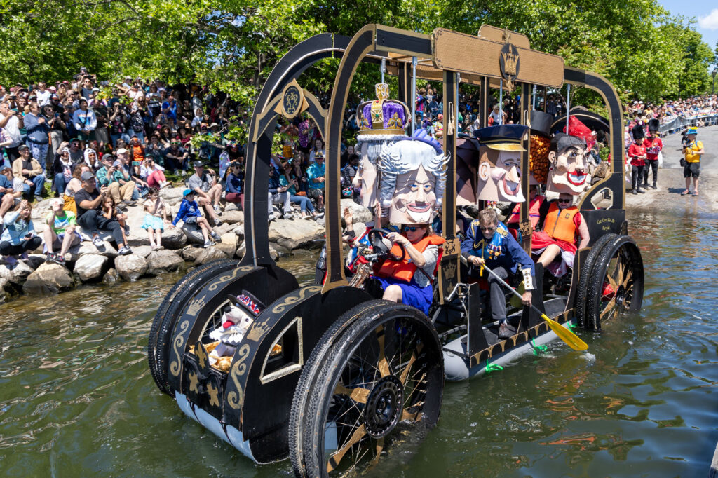 A floating vehicle made from cardboard floats on semi-hidden gray tanks. A crowd on the banks of the water watch as 6 participants wearing life jackets and oversized cartoon heads paddle and steer their way into the water.