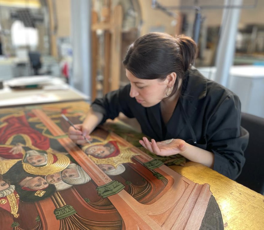Daniela holds a fine point paintbrush over a large late Medieval or early Renaissance-era European painting with a gold background on a horizontal work surface.